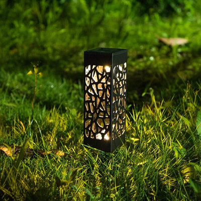 A vintage-style garden light with a black, ornate design, standing in a beautifully landscaped garden. The solar panel is visible on top, showcasing its ability to charge during the day. The light is illuminated, casting a warm glow on the surrounding plants and pathway, creating a cozy and inviting atmosphere.